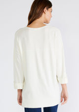 [Color: Cream] A back facing image of a brunette model wearing an ivory lightweight sweater tunic top. With three quarter length sleeves, a button roll tab, a v neckline, exposed seam details, a long tunic length, and side vents.