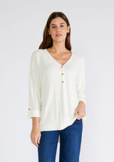 [Color: Cream] A front facing image of a brunette model wearing an ivory lightweight sweater tunic top. With three quarter length sleeves, a button roll tab, a v neckline, exposed seam details, a long tunic length, and side vents.
