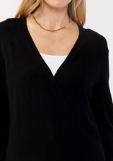 [Color: Black] A detail image of a blonde model wearing a black solid knit sweater with a wrap front and side button closures. A soft versatile sweater, perfect for spring styling. 