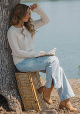[Color: Blush] A sitting image of a blonde model at the lake wearing a fuzzy open knit pullover sweater with a crew neckline, long sleeves, and a flutter cuff. Lightweight and breezy for the fall season. 