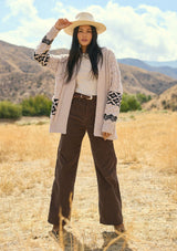 [Color: Natural/Black] A front facing image of a brunette model outside wearing a marled cable knit cardigan with a trendy black diamond pattern, an open front, and ribbed border. A southwestern inspired knit cardigan with a bohemian touch. 