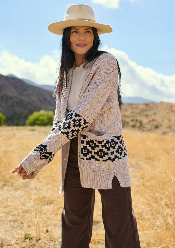 [Color: Natural/Black] A front facing image of a brunette model outside wearing a marled cable knit cardigan with a trendy black diamond pattern, an open front, and ribbed border. A southwestern inspired knit cardigan with a bohemian touch. 
