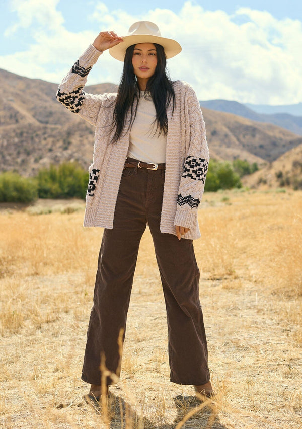 [Color: Natural/Black] A front facing image of a brunette model outside wearing a marled cable knit cardigan with a trendy black diamond pattern, an open front, and ribbed border. A southwestern inspired knit cardigan with a bohemian touch. 