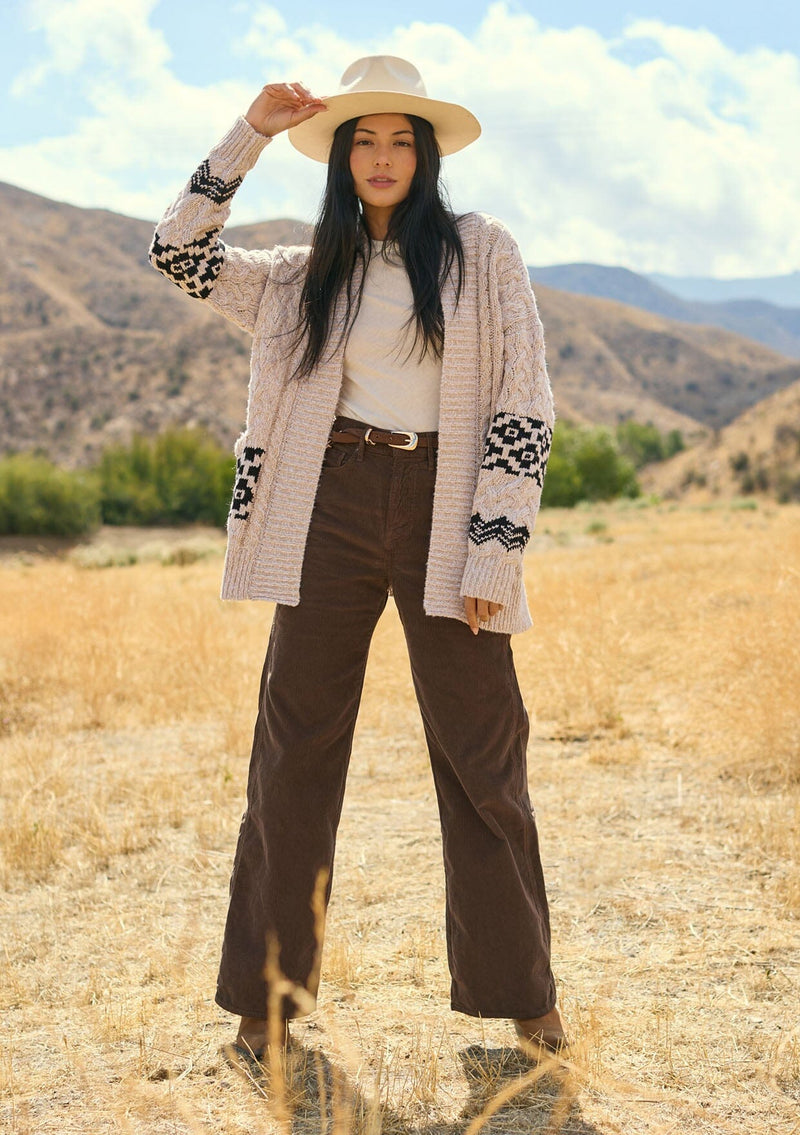 [Color: Natural/Black] A front facing image of a brunette model outside wearing a marled cable knit cardigan with a trendy black diamond pattern, an open front, and ribbed border. A southwestern inspired knit cardigan with a bohemian touch. 