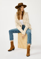 [Color: Natural/Dusty Pink] A sitting image of a blonde model wearing a beige southwestern boho sweater jacket with a zipper front and ribbed trims. A fall cozy sweater. 