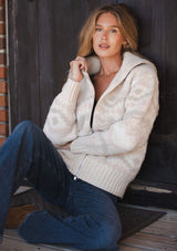 [Color: Natural/Dusty Pink] A sitting image of a blonde model wearing a beige southwestern boho sweater jacket with a zipper front and ribbed trims. A fall cozy sweater. 