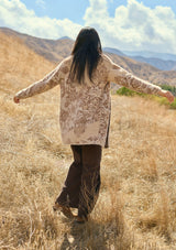 [Color: Sand] A back facing image of a brunette model outside wearing a brown floral print cardigan with a soft, fuzzy texture featuring an open front and patch pockets. A fall bohemian cardigan for casual styling. 