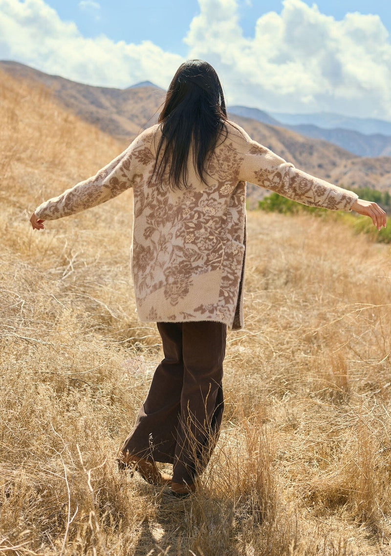 [Color: Sand] A back facing image of a brunette model outside wearing a brown floral print cardigan with a soft, fuzzy texture featuring an open front and patch pockets. A fall bohemian cardigan for casual styling. 