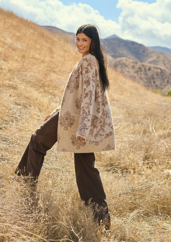 [Color: Sand] A side facing image of a brunette model outside wearing a brown floral print cardigan with a soft, fuzzy texture featuring an open front and patch pockets. A fall bohemian cardigan for casual styling. 