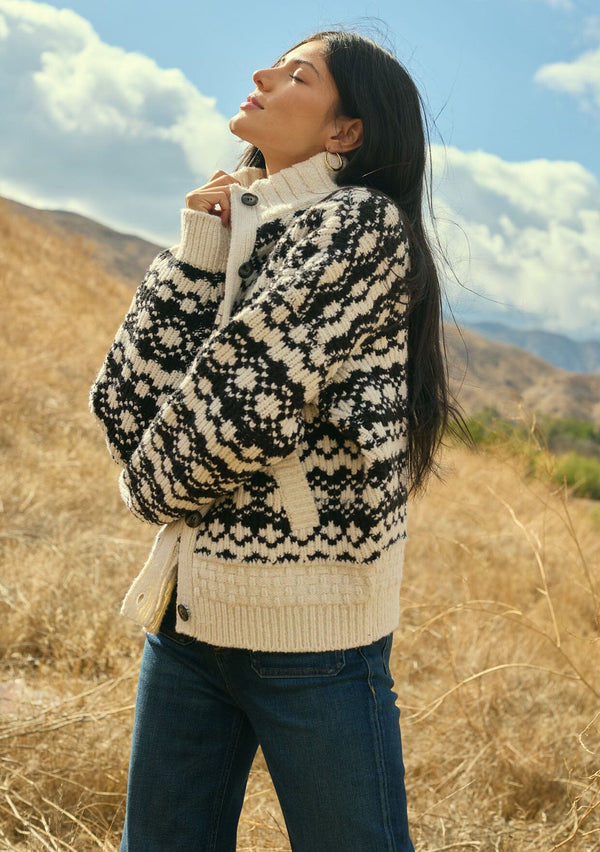 [Color: Cream/Black] A side facing image of a brunette model wearing a black and cream button front knit sweater with a high neckline and side pockets. A bohemian knit sweater for cozy fall styling. 