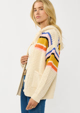 [Color: Natural/Mustard] A side facing image of a blonde model wearing a cream knit cardigan with a colorful striped pattern. Laid back with a hoodie, patch pockets, and button front. A cool girl fall essential!