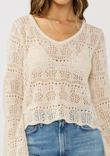 [Color: Natural] A detail image of a brunette model wearing a beige crochet knit top with long bell sleeves and a v-neckline. A breezy beach top for spring and summer. 