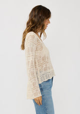 [Color: Natural] A side facing image of a brunette model wearing a beige crochet knit top with long bell sleeves and a v-neckline. A breezy beach top for spring and summer. 