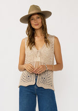 [Color: Natural] A front facing image of a brunette model wearing a beige crochet knit vest with a tie front. 