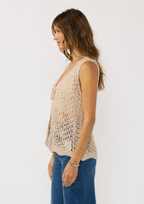 [Color: Natural] A side facing image of a brunette model wearing a beige crochet knit vest with a tie front. 