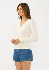 [Color: Natural] A side facing image of a brunette model wearing an off white waffle knit sweater top with a v neckline and a button front detail. 