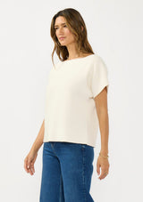[Color: Natural] A side facing image of a brunette model wearing a white sweater top with short sleeves and a boat neckline. 