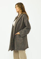 [Color: Dark Heather Brown] A side facing image of a brunette model wearing a brown cozy wool blend coatigan with an oversized fit, hoodie, and side pockets. 