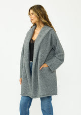 [Color: Heather Charcoal] A side facing image of a brunette model wearing a grey cozy wool blend coatigan with an oversized fit, hoodie, and side pockets. 
