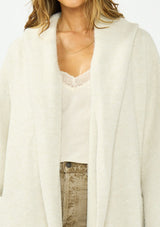 [Color: Heather Cream] A detail image of a brunette model wearing a cream cozy wool blend coatigan with an oversized fit, hoodie, and side pockets. 