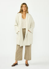 [Color: Heather Cream] A front facing image of a brunette model wearing a cream cozy wool blend coatigan with an oversized fit, hoodie, and side pockets. 