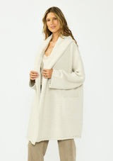 [Color: Heather Cream] A side facing image of a brunette model wearing a cream cozy wool blend coatigan with an oversized fit, hoodie, and side pockets. 