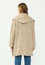 [Color: Heather Praline] A back facing image of a brunette model wearing a beige cozy wool blend coatigan with an oversized fit, hoodie, and side pockets. 