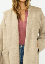 [Color: Heather Praline] A detail image of a brunette model wearing a beige cozy wool blend coatigan with an oversized fit, hoodie, and side pockets. 