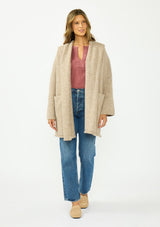 [Color: Heather Praline] A front facing image of a brunette model wearing a beige cozy wool blend coatigan with an oversized fit, hoodie, and side pockets. 
