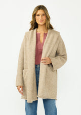[Color: Heather Praline] A front facing image of a brunette model wearing a beige cozy wool blend coatigan with an oversized fit, hoodie, and side pockets. 