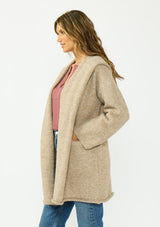 [Color: Heather Praline] A side facing image of a brunette model wearing a beige cozy wool blend coatigan with an oversized fit, hoodie, and side pockets. 