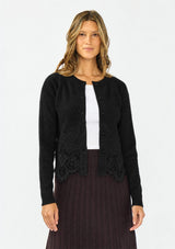 [Color: Black] A front facing image of a brunette model wearing a black lace cardigan with a button front. Paired with  a white top and a brown ribbed skirt. 