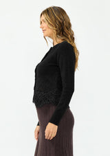 [Color: Black] A side facing image of a brunette model wearing a black lace cardigan with a button front. Paired with  a white top and a brown ribbed skirt.