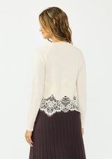 [Color: Bone] A back facing image of a brunette model wearing an ivory lace cardigan with a button front. Paired with  a white top and a brown ribbed skirt. 