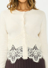 [Color: Bone] A detail image of a brunette model wearing an ivory lace cardigan with a button front. Paired with a brown ribbed skirt. 