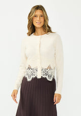 [Color: Bone] A front facing image of a brunette model wearing an ivory lace cardigan with a button front. Paired with  a white top and a brown ribbed skirt. 