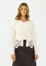 [Color: Bone] A front facing image of a brunette model wearing an ivory lace cardigan with a button front. Paired with  a white top and a brown ribbed skirt. 