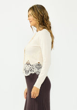 [Color: Bone] A side facing image of a brunette model wearing an ivory lace cardigan with a button front. Paired with  a white top and a brown ribbed skirt. 
