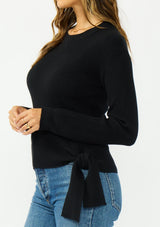 [Color: Black] A side facing image of a brunette model wearing a waffle knit sweater with a crew neckline and a side tie detail. 