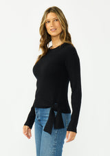 [Color: Black] A side facing image of a brunette model wearing a waffle knit sweater with a crew neckline and a side tie detail. 