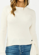 [Color: Natural] A detail image of a brunette model wearing an off white waffle knit sweater with a crew neckline and a side tie detail. 