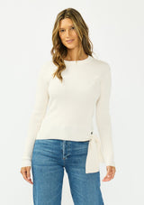 [Color: Natural] A front facing image of a brunette model wearing an off white waffle knit sweater with a crew neckline and a side tie detail. 