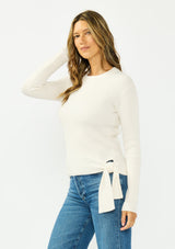 [Color: Natural] A side facing image of a brunette model wearing an off white waffle knit sweater with a crew neckline and a side tie detail. 