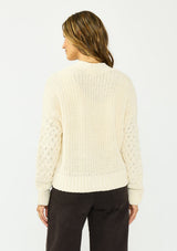 [Color: Natural] A back facing image of a brunette model wearing a cream knit cardigan with a button front and v neckline. Paired with dark brown denim. 