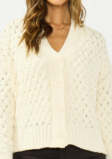 [Color: Natural] A detail image of a brunette model wearing a cream knit cardigan with a button front and v neckline. Paired with dark brown denim. 
