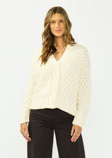 [Color: Natural] A front facing image of a brunette model wearing a cream knit cardigan with a button front and v neckline. Paired with dark brown denim. 
