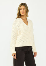 [Color: Natural] A side facing image of a brunette model wearing a cream knit cardigan with a button front and v neckline. Paired with dark brown denim. 