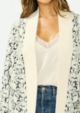 [Color: Cream/Black] A detail image of a brunette model wearing a snake print cardigan with a fuzzy texture, an open front, and contrast trim. Paired with a lace camisole and a vintage dark wash denim. 