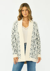 [Color: Cream/Black] A front facing image of a brunette model wearing a snake print cardigan with a fuzzy texture, an open front, and contrast trim. Paired with a lace camisole and a vintage dark wash denim. 
