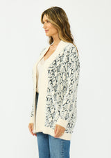 [Color: Cream/Black] A side facing image of a brunette model wearing a snake print cardigan with a fuzzy texture, an open front, and contrast trim. Paired with a lace camisole and a vintage dark wash denim. 